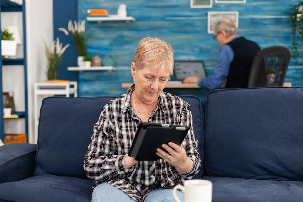 Pensioner lady browsing on internet sitting on couch. Elderly woman using moder technoloy tablet pc in home living room and husband working on laptop computer